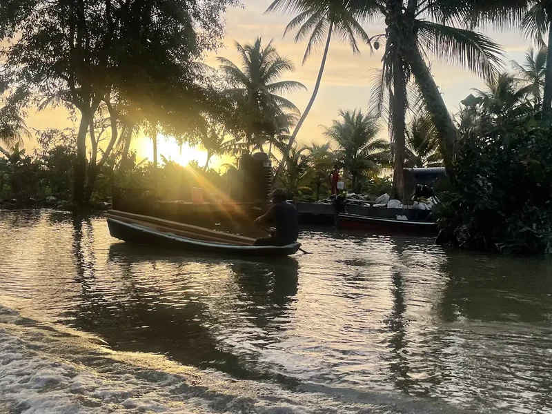 A small boat passes palms and riverside buildings at sunset.