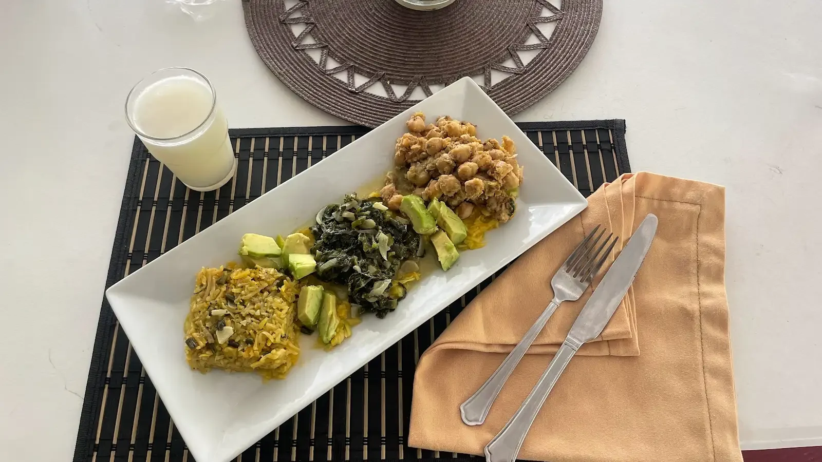 A rectangular plate with several cooked dishes and vegetables beside cutlery and a drink.