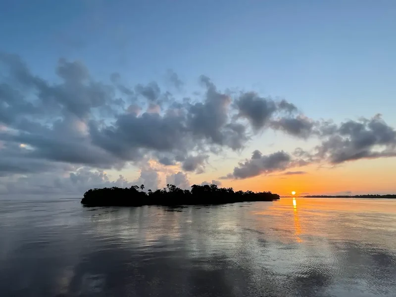 Blue and orange evening sky reflected in calm water around a small wooded island.
